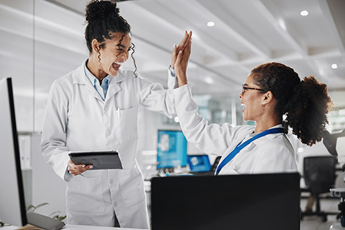 Two female scientists in a lab setting, both wearing white lab coats and glasses, exchanging a celebratory high-five. One holds a tablet, while they smile in a bright, modern laboratory. Below the image is a green banner with white text that reads 'By Scientists, For Scientists'