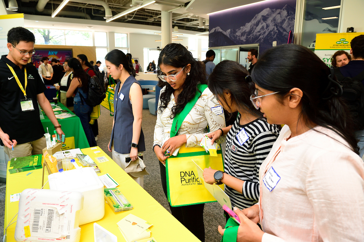 Students interact with representatives at a Zymo Research booth, exploring sample preparation products displayed on a bright yellow table.
