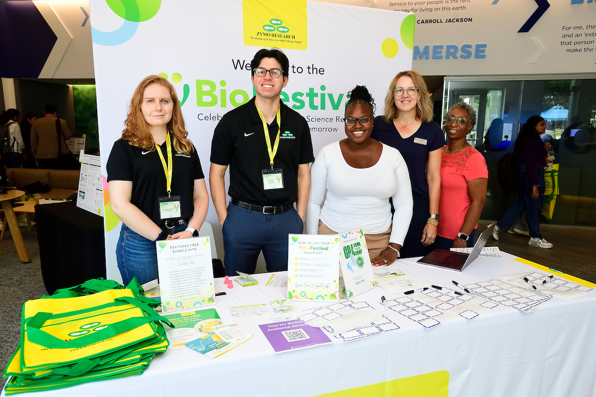 Zymo Research representatives smile behind their BioFestival booth featuring informational materials, green and yellow tote bags, and sample kit flyers.