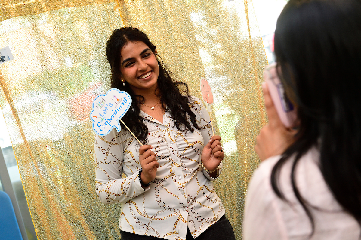 A woman smiles while posing for a photo with playful science-themed props, including one that says “Let’s Experiment,” in front of a glittery gold backdrop.