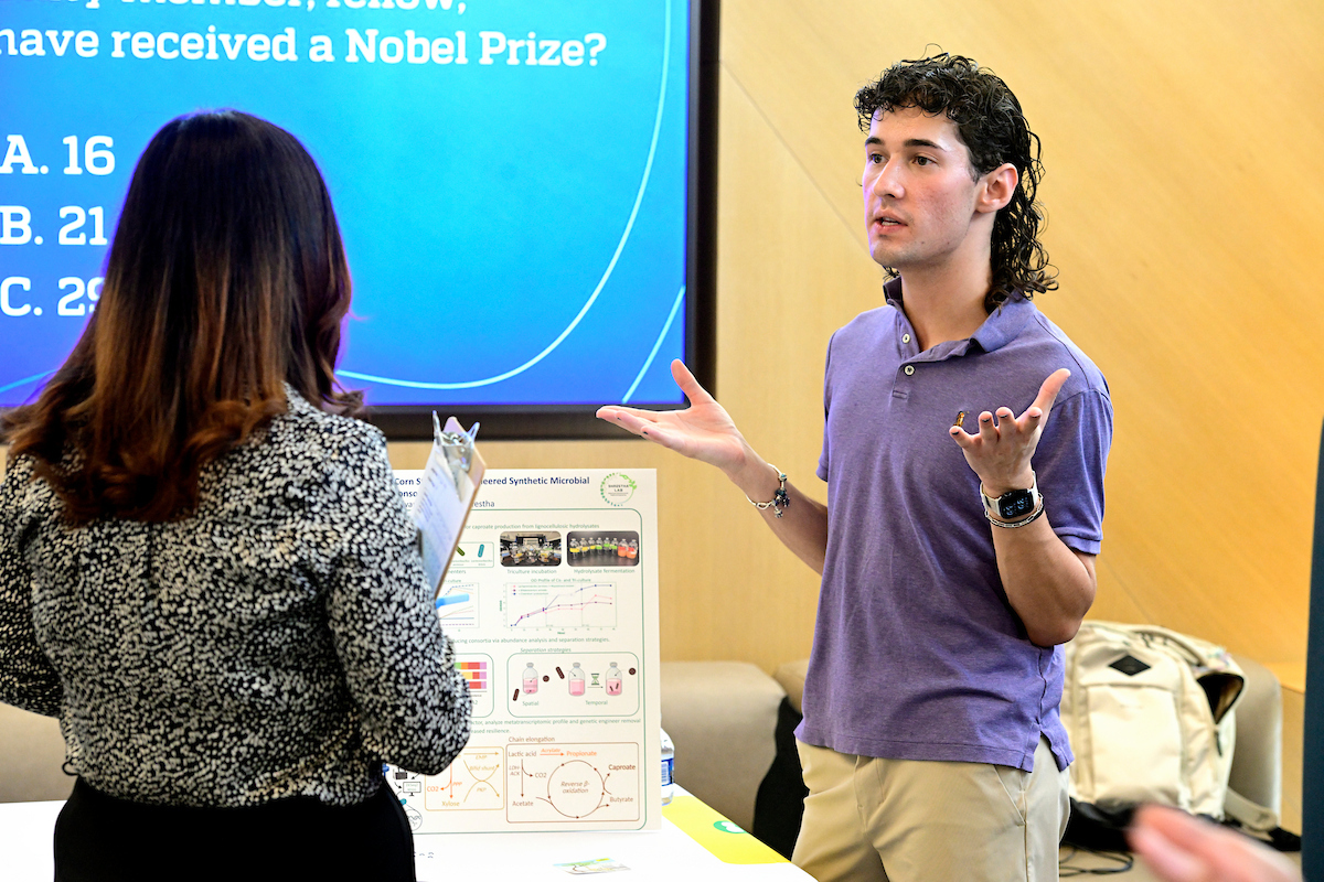 A student in a purple shirt presents a scientific poster to a judge holding a clipboard during a BioFestival event, with a quiz question projected on a screen behind them.
