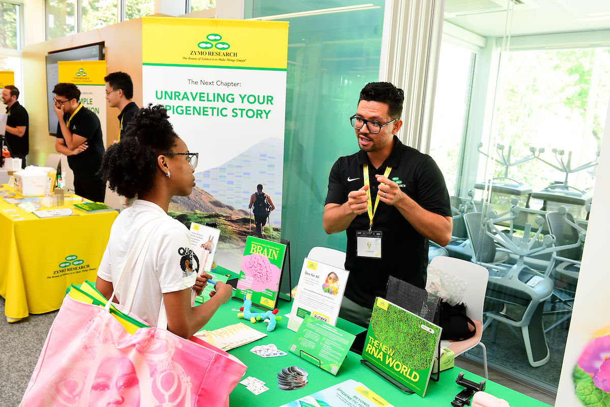 A Zymo Research representative talks with an attendee at a booth displaying educational materials about epigenetics and RNA research during a science event.