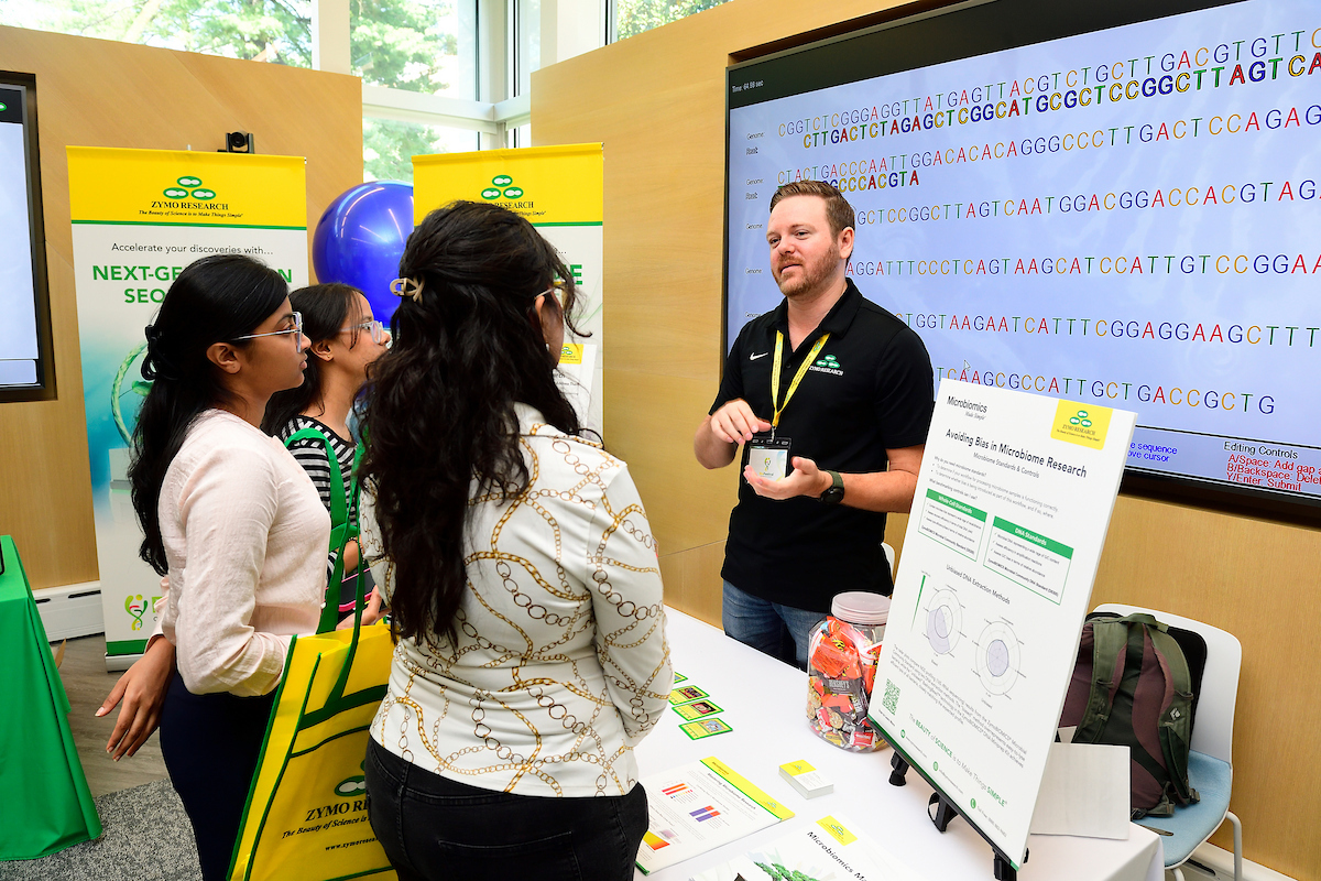A Zymo Research representative explains microbiome research methods to attendees at a booth featuring educational displays and sequencing visuals during the BioFestival event.
