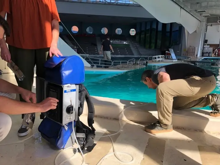 Two individuals are interacting near a dolphin tank. One person, in an orange shirt, is holding a piece of equipment while the other, crouched down and wearing a black shirt, inspects the tank's edge. A blue backpack with attached equipment sits on the ground beside them