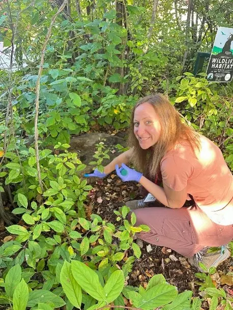 A woman with long hair, wearing gloves and casual clothing, is kneeling beside a rock in a lush area filled with plants. She is smiling while working on a task among the greenery. A sign in the background indicates it is a wildlife habitat