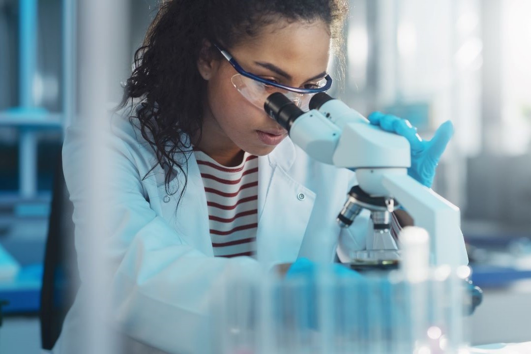A scientist wearing safety goggles, gloves, and a lab coat closely examines a sample through a microscope in a laboratory setting